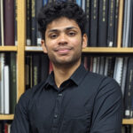 A headshot photo of Ashwin standing in front of bookshelves.
